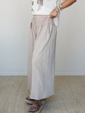 A side-angle shot of a wide-leg maxi pant in washed taupe, styled with a cropped graphic tee and espadrille platform sandals — the kind of effortless outfit that looks intentional without trying.