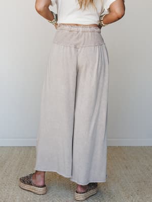 A back-facing shot of wide-leg taupe pants with a smocked waistband, styled with a cropped white tee, beaded bracelets, and leopard print espadrille flatforms — effortless and a little unexpected.
