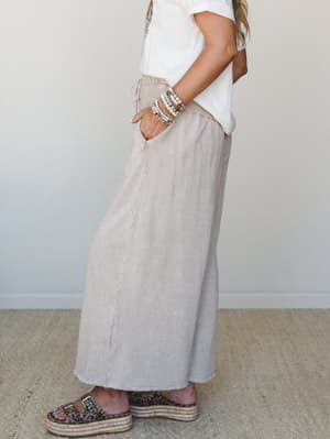 A side-profile shot of a wide-leg pant in washed taupe, styled with a white tee, stacked bead bracelets, and leopard print platform sandals — the kind of effortless outfit that looks like you tried just enough.