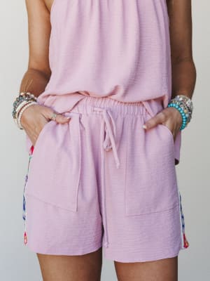 A close mid-shot of a lavender-pink matching set — relaxed shorts with a drawstring waist and side floral trim peeking through — styled with stacks of beaded and crystal bracelets that feel collected, not coordinated.