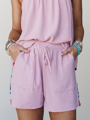 A close mid-shot of a lavender-pink matching set — relaxed shorts with a drawstring waist and side floral trim peeking through — styled with stacks of beaded and crystal bracelets that feel collected, not coordinated.