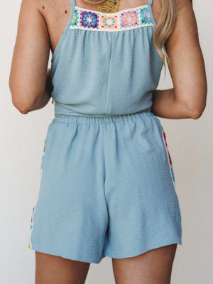 A back-view shot of a light blue romper with colorful granny square crochet detailing along the neckline and side seams — the kind of piece that looks like it was made for a slow summer morning somewhere warm.