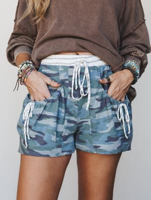 A mid-shot of blue-toned camo drawstring shorts styled with an oversized washed brown sweatshirt and stacked boho bracelets — relaxed, lived-in, and the kind of outfit you throw on and somehow look put-together.