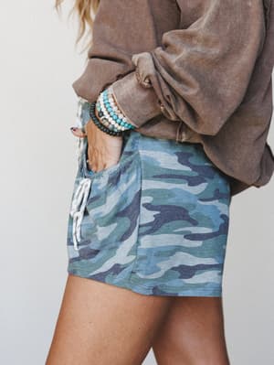 A close-up lifestyle shot of blue-toned camo drawstring shorts styled with a washed brown oversized sweatshirt and a stack of beaded bracelets — casual and collected, like a Saturday with nowhere to be.