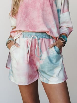 A tie-dye matching set — oversized top and drawstring shorts in cotton candy pink and sky blue — styled with stacks of boho bracelets, hands tucked into pockets like she's perfectly comfortable with being this cute.