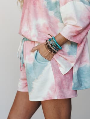 A close-up of a matching tie-dye set — oversized top with a side slit and drawstring shorts — styled with a stack of beaded bracelets, hand tucked casually into a pocket.