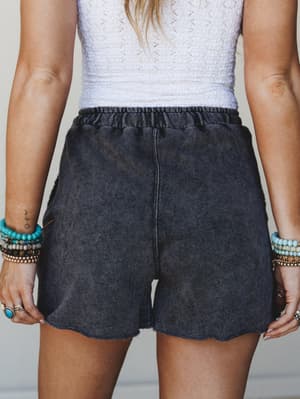 A close-up mid-shot of acid wash charcoal shorts with a relaxed fit and raw hem, styled with a white crochet top and stacked boho bracelets — the kind of effortless summer outfit that looks like it came together without trying.