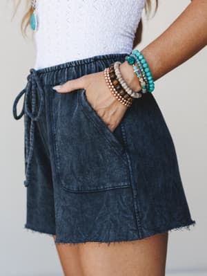 A close-up mid-shot of acid wash navy shorts with a drawstring waist and raw hem, styled with a white textured top and a stack of boho bead bracelets — the kind of effortless summer outfit that looks like it took zero effort and exactly the right amo