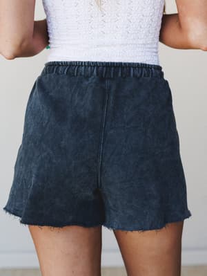 A close back-view shot of acid-washed navy pull-on shorts with a raw hem, paired with a white textured top — the kind of effortless combo that makes a casual day feel intentional.