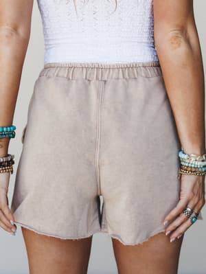 A close-up torso shot of washed taupe fleece shorts with a raw hem paired with a white smocked top, styled with stacked beaded and silver bracelets for a laid-back boho feel.