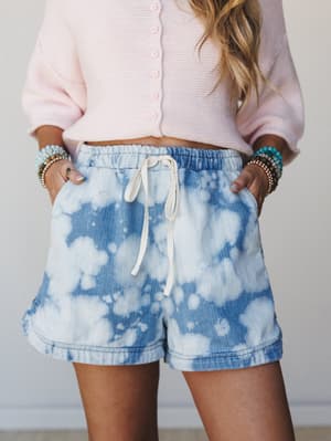 A close mid-shot of bleach tie-dye denim shorts with a soft drawstring waist, styled with a blush pink cropped cardigan and layered beaded bracelets — casual and effortlessly put-together.