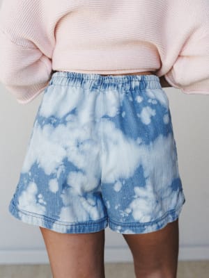 A close-up back view of bleach tie-dye denim shorts with an elastic waistband, styled with a soft pink cropped knit sweater — casual and expressive in the best way.