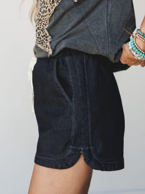 A close-up mid-shot of dark denim shorts styled with a charcoal graphic tee, leopard print top layer, and a stack of turquoise beaded bracelets — casual and collected with just enough personality.