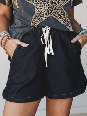A close-up mid-shot of dark denim drawstring shorts styled with a washed graphic tee and stacked boho bracelets — effortless and a little lived-in, the kind of outfit you'd actually reach for on repeat.
