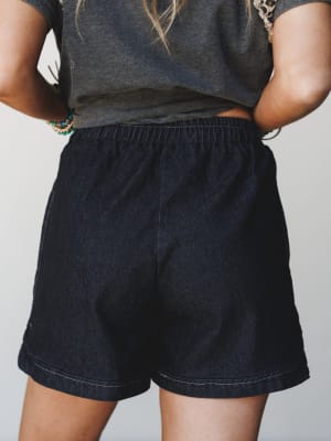 A close-up back view of relaxed-fit dark denim shorts with an elastic waistband, styled with a charcoal graphic tee and stacked beaded bracelets — the kind of effortless summer outfit that looks like you tried just enough.