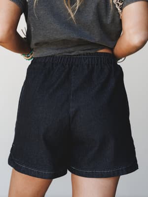 A close-up back view of relaxed-fit dark denim shorts with an elastic waistband, styled with a charcoal graphic tee and stacked beaded bracelets — the kind of effortless summer outfit that looks like you tried just enough.