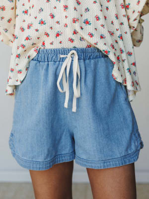 A mid-shot of soft denim pull-on shorts with a cream drawstring waist, styled with a floral print long-sleeve top — the kind of easygoing outfit that makes a Tuesday feel worth getting dressed for.