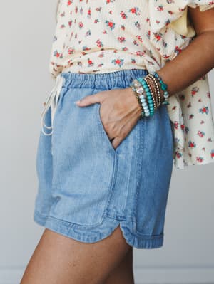 A close-up waist-to-thigh shot of light wash denim shorts paired with a floral ruffle top and a stacked bracelet set — the kind of effortless summer outfit that somehow looks like you tried just enough.