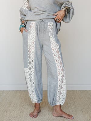 A heather grey jogger pant with cream lace side panels runs full-length on a barefoot model styled with stacked turquoise and stone bracelets — cozy-meets-boho in the most wearable way.