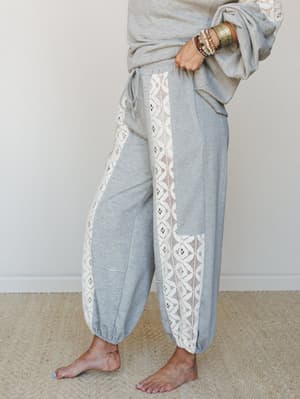 A close-up side view of heathered grey jogger pants with wide lace side panels running the full length of the leg, styled barefoot with stacked bracelets and a matching grey sweatshirt — cozy and expressive all at once.
