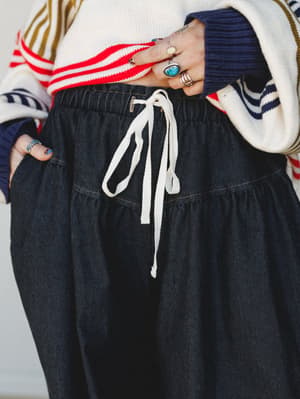 A close-up waist shot of a dark denim wide-leg pant with a cream drawstring tie and tiered seam detail, styled with a striped varsity-style sweater and layered silver rings — casual and collected with a bit of personality.