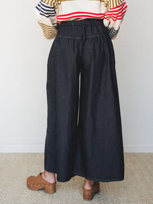 Shot from behind, a model wears dramatically wide-leg dark denim pants with an elastic gathered waist, styled with a multicolor stripe sweater and brown clogs — the kind of outfit that looks effortless but clearly wasn't an accident.