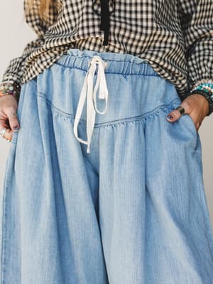 A close-up detail shot of flowy light denim wide-leg pants with a gathered drawstring waist, styled with a black-and-cream gingham top and layered beaded bracelets — easy and expressive, the kind of outfit that feels as good as it looks.