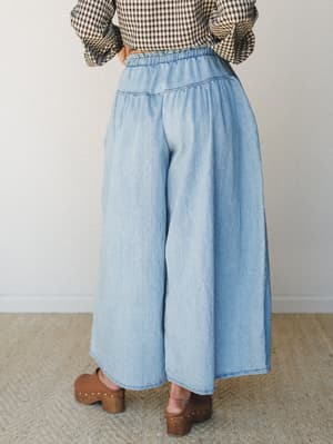Shot from behind, this look pairs ultra-wide-leg chambray pants in a soft light denim wash with a black-and-white gingham top and warm brown clogs — relaxed and intentional, like she just knows.