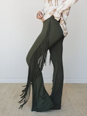 A side-profile shot of olive flare pants with dramatic long fringe running down the outer seam, styled with a cream novelty-print sweater — the kind of outfit that makes a statement without trying too hard.