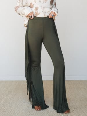 Olive flare pants with dramatic full-length side fringe, styled with a western-print sweater, turquoise rings, and studded clogs — the kind of outfit that moves when you do.