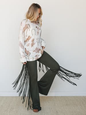 A boho-western outfit pairing olive fringe flare pants with dramatic side fringe against a model in a cream novelty-print sweatshirt — the kind of look that makes a statement without trying too hard.