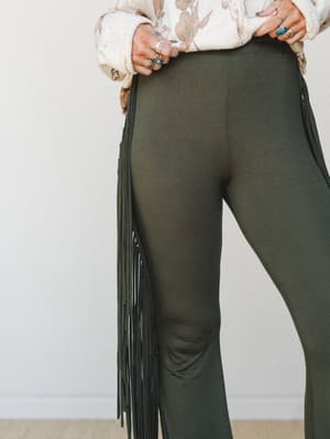A close-up detail shot of olive flare pants with long side fringe trim, styled with a cream floral sweater and stacked silver and turquoise rings — earthy, free-spirited, and unapologetically boho.