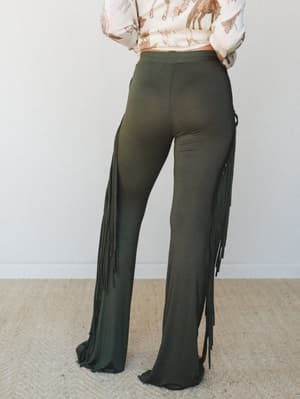 A back-view shot of olive wide-leg flare pants with dramatic long side fringe, styled with a cream horse-print top — the kind of outfit that makes an entrance without trying.