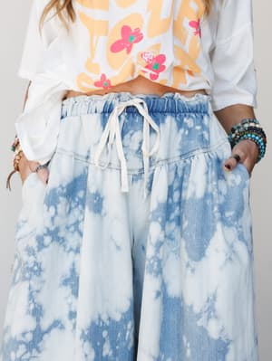 A close-up mid-shot of wide-leg bleach-dyed denim pants with a relaxed drawstring waist, styled with a cropped graphic tee and stacked beaded bracelets — the kind of outfit that looks effortless but is clearly doing a lot.