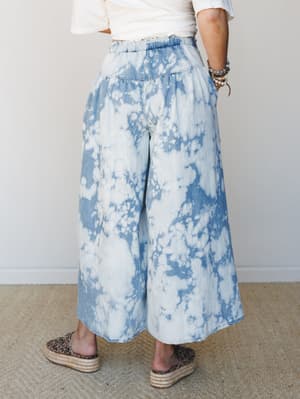 A back-view shot of wide-leg bleach-dye denim pants with a paperbag waist, styled with a white crop top, espadrille platform sandals, and stacked bead bracelets — easy, expressive, and very much a summer Saturday.