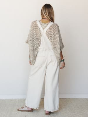 Shot from the back, a model layers an open-knit poncho over wide-leg ivory overalls, styled with stacked turquoise bracelets and strappy sandals — the kind of effortless, collected outfit that looks like it came together naturally.
