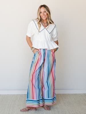 A smiling model pairs a white puff-sleeve sailor-collar top with bold multicolor striped wide-leg pants — the kind of outfit that makes a grocery run feel like a vacation.