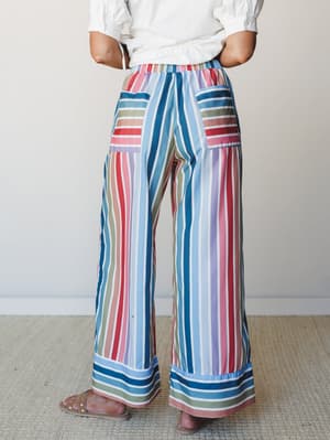 Wide-leg striped pants in a cheerful multi-color palette styled with a white top and tan sandals, shot from behind against a soft neutral interior — the kind of pants that do all the talking.