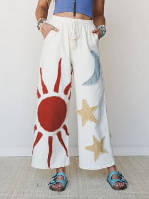 Wide-leg ivory pants with raw-edge appliqué sun, moon, and star motifs steal the show here — styled with a periwinkle crop top, turquoise sandals, and stacked bracelets for a free-spirited, celestial-meets-summer look.