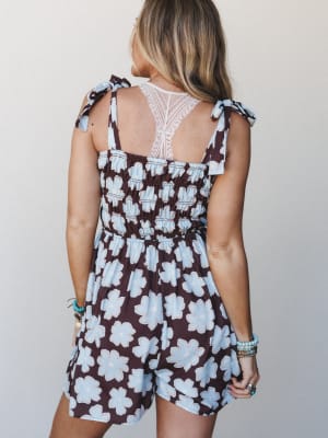 A back view of a floral romper in deep brown and powder blue, showing off the smocked bodice, lace back panel, and tie shoulder straps — styled with stacked turquoise bracelets for an effortlessly boho feel.