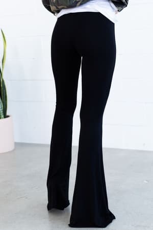 Wearing the Cher Solid Raw Edge Flare Pant in Black, a flared-leg style crafted from stretchy fabric for comfort and style all day. Shown indoors with a jacket, on a concrete floor beside a white wall and potted snake plant.