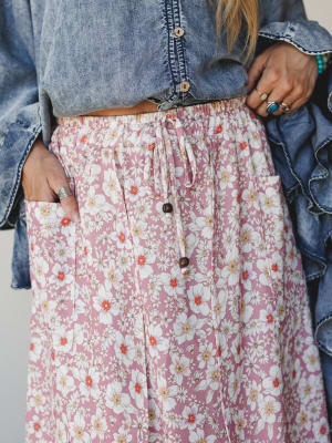 A close-up detail shot of a flowy floral midi skirt in dusty rose, styled with a cropped denim top — the button-front placket, drawstring waist, and patch pocket are front and center, with silver and turquoise rings adding that effortless boho finish