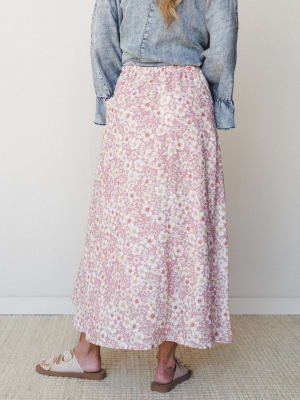 A back-view shot of a flowy maxi skirt in dusty rose with an all-over white daisy print, styled with a washed denim top and neutral leather sandals against a clean studio wall.