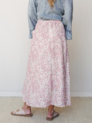 A back-view shot of a flowy maxi skirt in dusty rose with an all-over white daisy print, styled with a washed denim top and neutral leather sandals against a clean studio wall.
