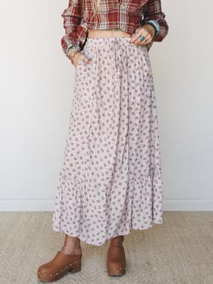 A flowy ditsy floral maxi skirt in soft blush-taupe pairs with a cropped red plaid shirt and wooden clogs — the kind of effortless boho layering that feels like it was thrown on in the best possible way.