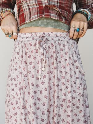 A close-up torso shot showing a ditsy floral maxi skirt in dusty blush-taupe layered under a cropped plaid top and ribbed cami, styled with stacked rings and beaded bracelets for a well-worn boho feel.