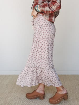 A side-profile shot of a flowy ditsy floral maxi skirt in cream and dusty rose, styled with a red plaid top, stacked boho bracelets, and studded wood clogs — the kind of outfit that feels equally at home on a farm stand run or a fall weekend away.