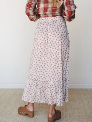 A back-view shot of a flowy ditsy floral maxi skirt in soft cream paired with a red plaid shirt and wood-studded clogs — the kind of lived-in mix that looks effortless but totally intentional.