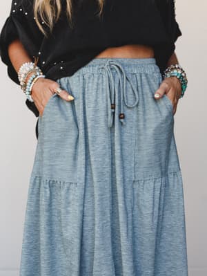 A close-up shot of a heather teal tiered maxi skirt with a drawstring waist, styled with a knotted black top and stacked boho bracelets — the kind of effortless outfit that looks intentional without trying.