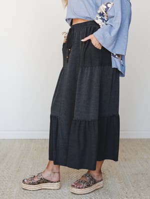 A charcoal two-tone tiered maxi skirt styled with a blue floral-sleeved top and leopard print platform sandals, shot from the side against a clean studio wall — relaxed and wearable with just enough personality.